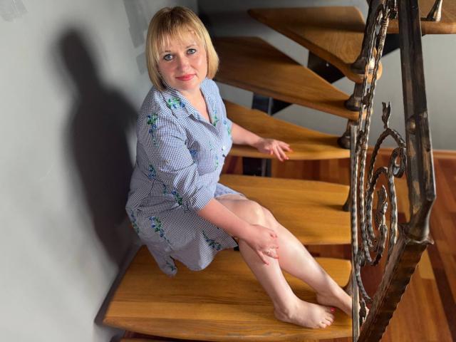 KiraSmiling with blonde bob, wearing a blue check dress with green floral patterns, sitting on wooden stairs, barefoot, smiling, hands on stairs, metal railing, shadow on wall.