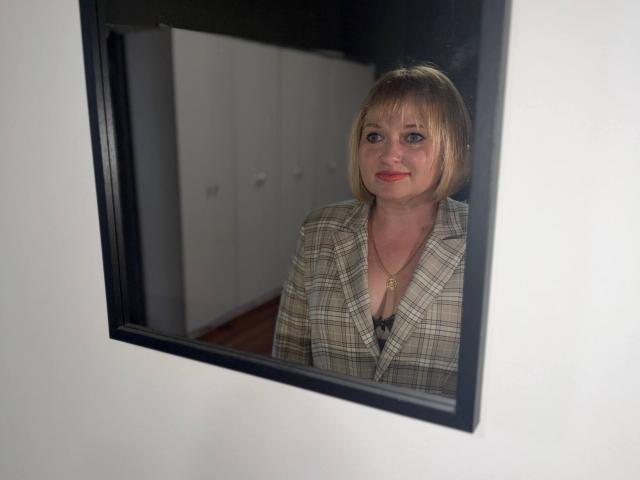 KiraSmiling heeft blond haar, rode lippenstift en draagt een geruit blazer met een gouden ketting, wat weergegeven wordt in een spiegel met een zwarte lijst. Haar borsten zijn licht zichtbaar onder de blazer..