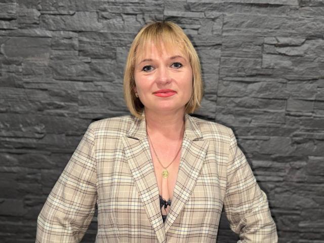 KiraSmiling has blonde bob, light skin, red lipstick, wearing plaid blazer, black bra visible, necklace with green pendant, standing in front of gray textured wall.