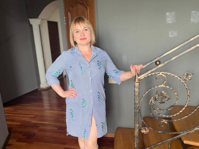 KiraSmiling stands in a blue dress with green floral patterns, holding a metal staircase railing with one hand, and has a slight smile, showing her pussy through a thigh-high slit.