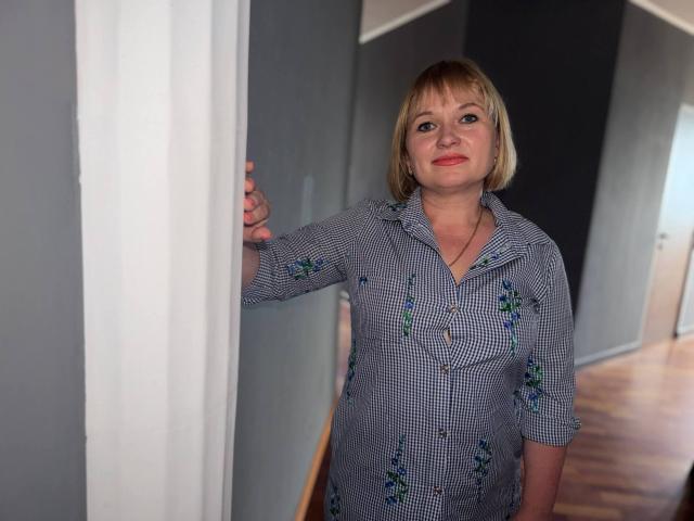 KiraSmiling, blonde woman with red lipstick, wearing a blue checkered shirt with green floral embroidery, standing in a doorway, holding a white curtain, wooden floor.