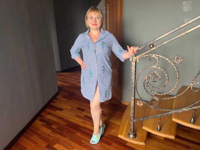 KiraSmiling with blonde hair, blue button-up dress, floral patterns, knee-length, blue slippers, standing by a metal staircase, wooden floor, gray wall, holding railing, smiling, red lipstick.