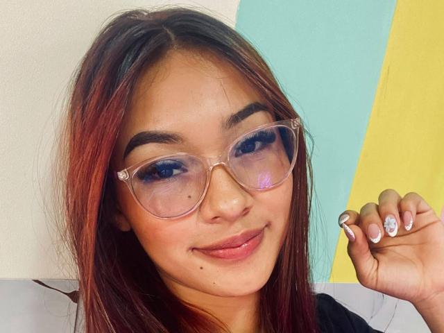 SashaHans has red-brown hair, wearing clear glasses, smiling with pinkish-blue eyeshadow, and showing off her manicured nails with white tips and a blue gem.