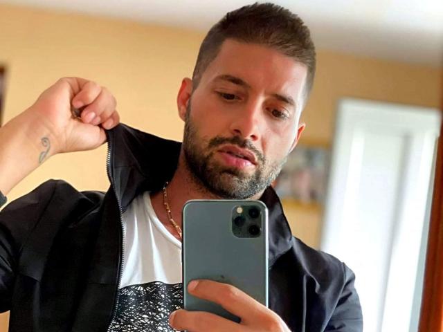 TommyDum with a short beard, brown hair, and a tattoo on his right forearm, taking a selfie in a mirror, wearing a black jacket over a white shirt.
