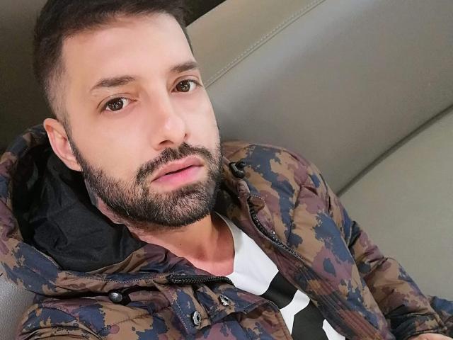 TommyDum with a trimmed beard and short brown hair, wearing a camo jacket over a white shirt, looking slightly down with a relaxed expression.