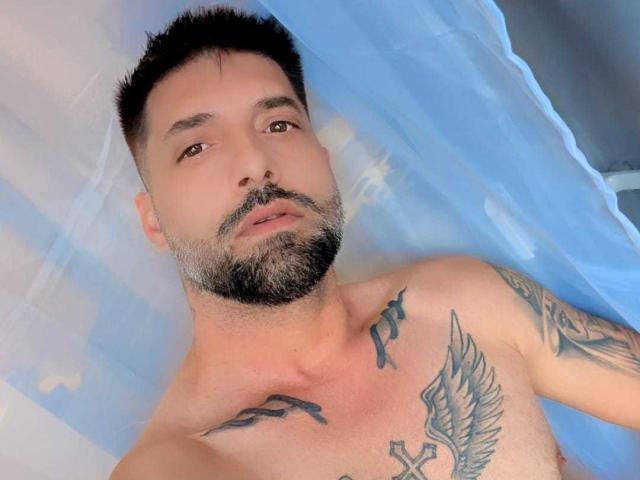TommyDum, shirtless with tattoos on chest and arms, has short black hair, beard, and brown eyes, lying on blue and white bed. His chest and tattoos are visible.