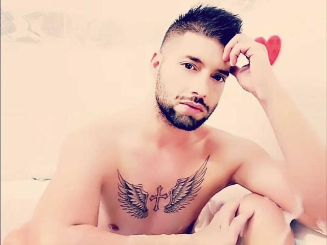 TommyDum, shirtless with a tattoo of wings and a cross on his chest, short dark hair, beard, looking at camera, hand on head, sitting on bed.