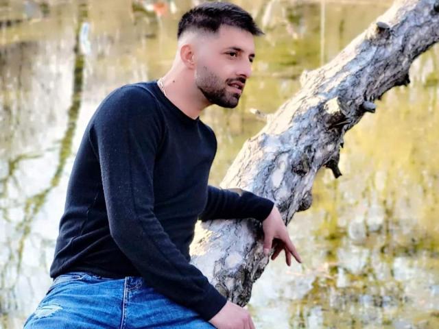 TommyDum with short dark hair and beard, wearing a black long-sleeve shirt and blue jeans, sitting on a tree branch, looking to the side with a serious expression.