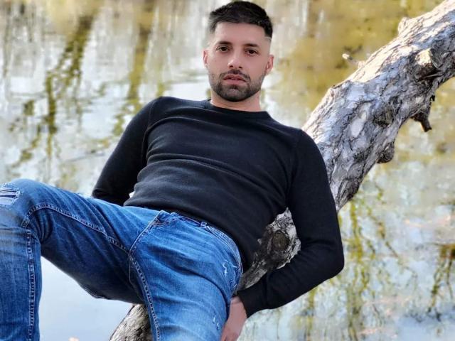 TommyDum, con una maglietta nera e jeans blu, appoggiato a un albero, gambe divaricate, espressione seria, capelli corti e scuri, leggera barba, mani in tasca, ambiente esterno informale..