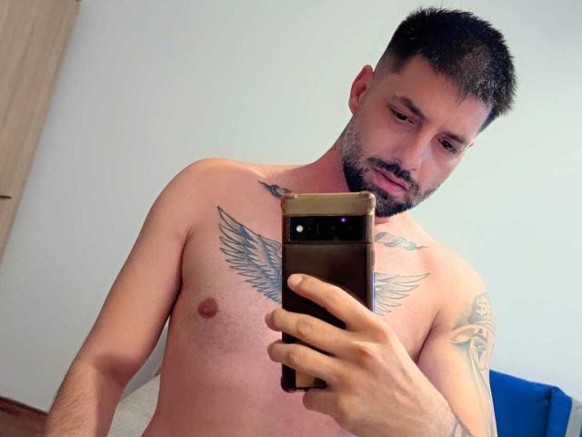 TommyDum, shirtless, with tattoos on chest and arm, holding phone, looking down, short dark hair, beard, fair skin, medium build, slightly muscular, taking a mirror selfie.