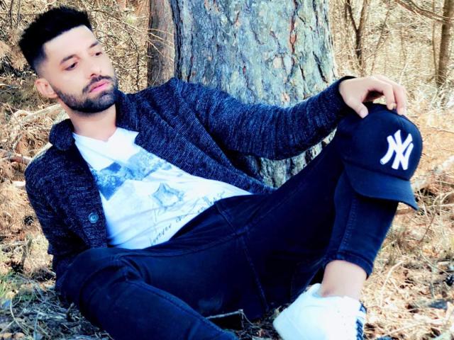 TommyDum with a black beard, wearing a blue cardigan, white graphic tee, black jeans, and a NY Yankees cap, sitting against a tree with one knee up, looking down.