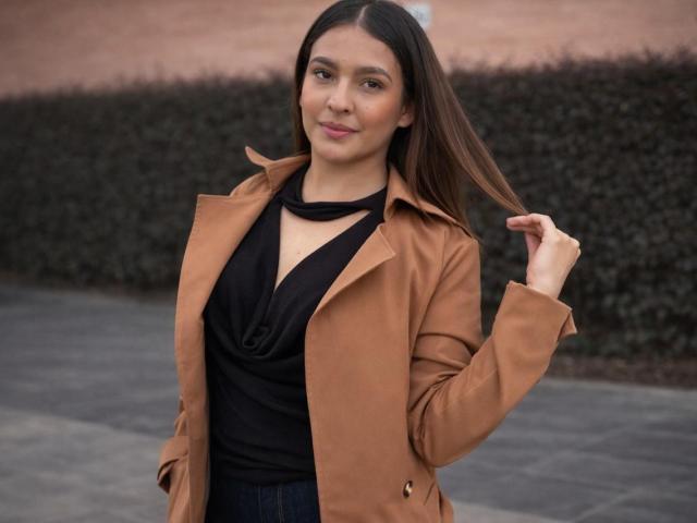 Talia Bonet, avec de longs cheveux bruns, portant un manteau beige par-dessus un haut noir, se tenant les cheveux, souriant doucement, debout sur un trottoir gris, et ayant une allure adorable..