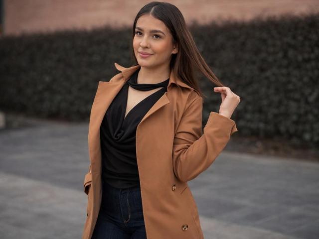 TaliaBonet, com cabelos castanhos longos, casaco bege, blusa preta e calças jeans escuras, sorrindo, segurando o cabelo, mostrando seios pequenos, em pé do lado de fora em um pavimento cinza..