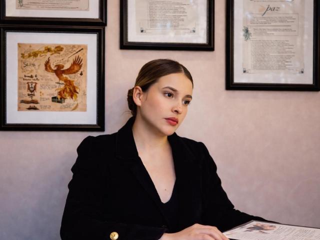 TaliaBonet, com cabelo castanho, usando um blazer preto, sentada, olhando para o lado, com as mãos em um documento, com obras de arte emolduradas e certificados em uma parede rosa ao fundo..