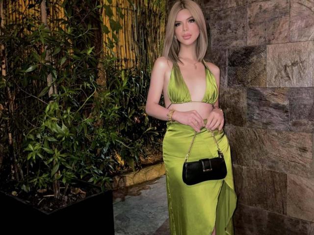 HotCumAdriana69x with blonde hair, green satin halter dress, black purse, holding it, showing tits, standing, holding arm, stone wall background, green bamboo.