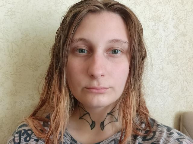 Charlesetta Lahr, con el cabello castaño-rubio y húmedo, ojos verdes y un tatuaje negro con forma de alas en el cuello. Lleva una camisa gris con estampado..