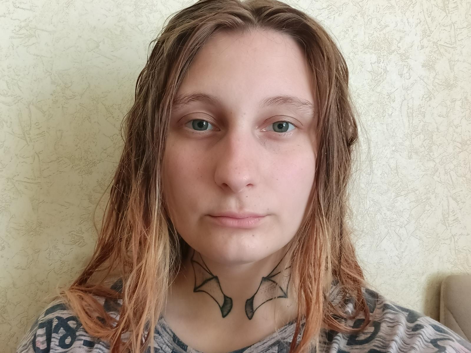Charlesetta Lahr, con el cabello castaño-rubio y húmedo, ojos verdes y un tatuaje negro con forma de alas en el cuello. Lleva una camisa gris con estampado..