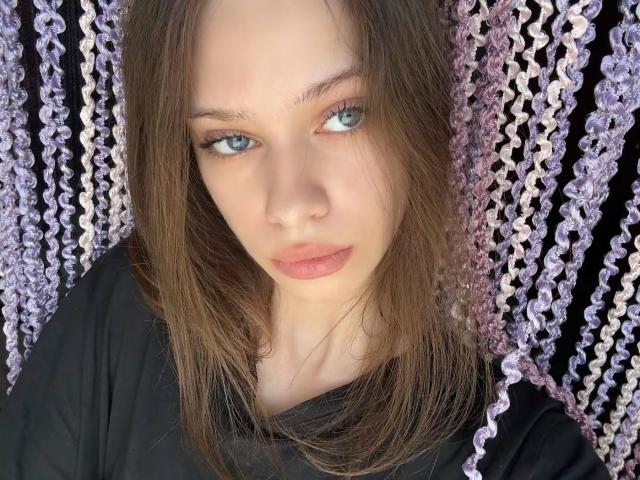 PreettyLaady tiene ojos azules, cabello castaño y liso, y un rostro neutro con labios ligeramente fruncidos. Lleva una camisa negra, y hay un fondo ondulado con un patrón púrpura..