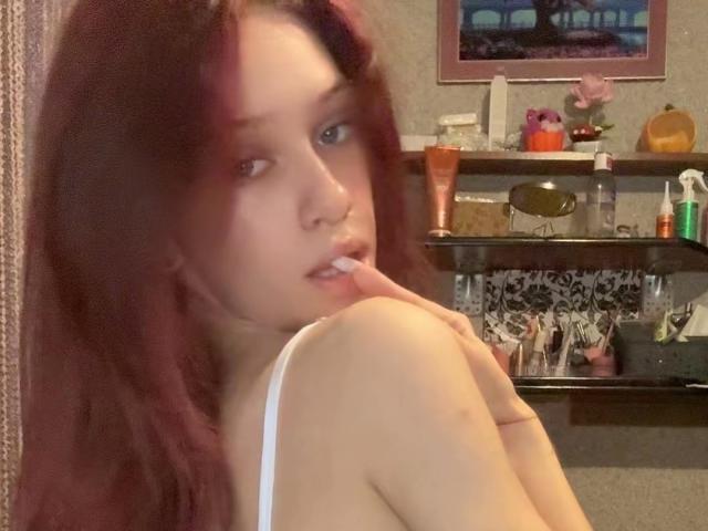 PreettyLaady avec des cheveux roux, une peau pâle et de petites seins, regardant par-dessus son épaule, mordant son doigt, portant un débardeur blanc, sur une photo de profil floue..