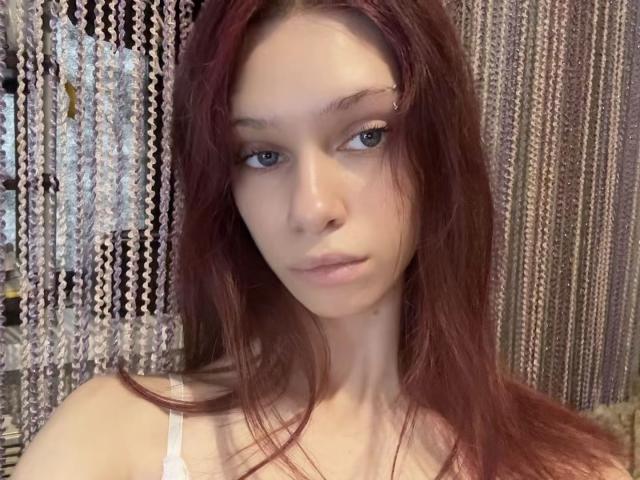 PreettyLaady ha lunghi capelli rossi, occhi azzurri e un'espressione neutra. Indossa una canottiera bianca che mette in mostra i suoi piccoli seni. L'immagine è un selfie..