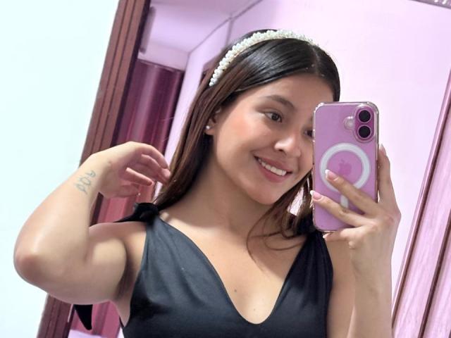 TaliaBonet mit braunen Haaren, die ein schwarzes Kleid und ein weißes Stirnband trägt, lächelt in ein Selfie vor einem Spiegel, hält ein rosa Handy und hebt ihren Arm, um ein Tattoo zu zeigen..