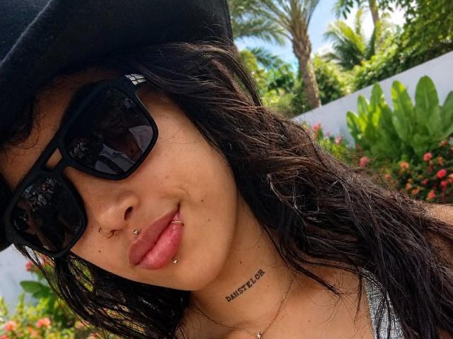 Krystal Evans, mit schwarzen Sonnenbrillen, einem schwarzen Hut und Nasenpiercings, hat ein Lippenpiercing und ein "LIEBE"-Tattoo, trägt eine silberne Halskette, hat welliges, dunkles Haar und rosafarbene Lippen..