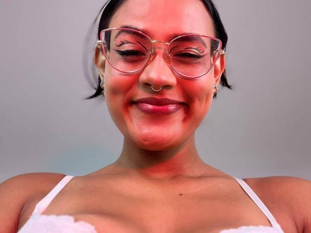 LilithRussoo has red skin, black hair, glasses, nose ring, earrings, white lace top, and a subtle smile. Her tits are small, and she's looking cute.