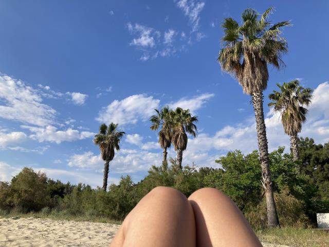 DeesseBleue's knees in the foreground, with tall palm trees and a blue sky with white clouds in the background. Her skin is tan, and the beach is sandy.