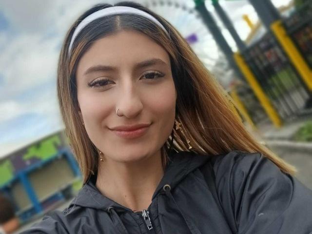 Kaiix tiene la piel de color marrón claro, cabello largo y castaño con una diadema blanca, lleva una sudadera negra, un pequeño aro en la nariz y aretes dorados, y está sonriendo ligeramente..