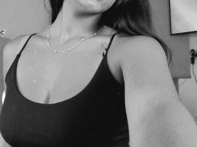 Selfie em preto e branco de SarahPrincess mostrando seus seios em uma regata listrada, com um colar e uma tatuagem no ombro direito, com a cabeça ligeiramente fora do quadro..