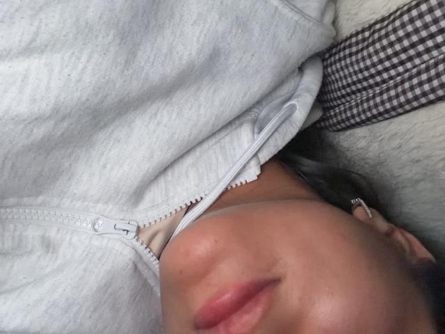 SarahPrincess's face, partially visible, with pink lips and a slight tan, wearing a gray zip-up shirt, partially unzipped, exposing a bit of white bra. Her ear with a stud.