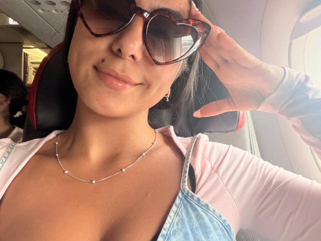SarahPrincess mit Schildpattbrille, lächelnd, trägt ein rosa Oberteil und ein blaues Jeanskleid, die Hand ist in der Nähe des Gesichts, sie trägt eine Halskette und Ohrringe, und sitzt auf einem Flugzeugsitz. Brüste sind unter dem Kleid sichtbar..