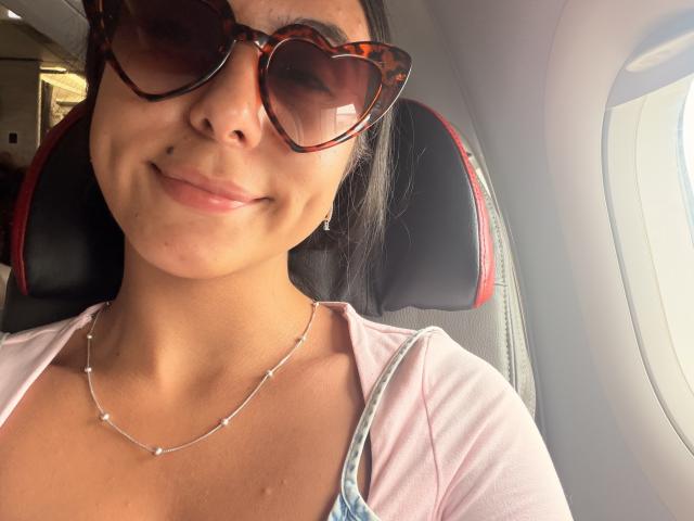SarahPrincess with dark brown eyes, wearing heart-shaped tortoiseshell sunglasses, a pink top, and a silver necklace, smiling in an airplane seat, showing her tits slightly.