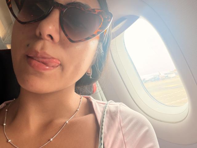 SarahPrincess with her tongue out, wearing tortoiseshell sunglasses, a pink top, and a bead necklace, has a small beauty mark above her left lip.