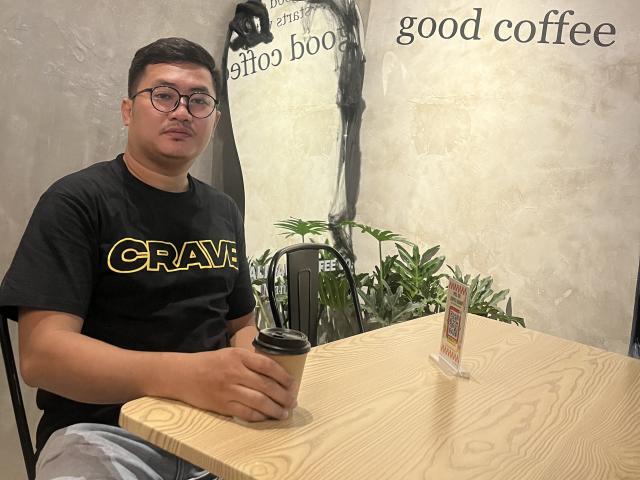 AkiCumshott, um homem corpulento de óculos, camisa preta, segurando uma xícara de café, sentado em uma mesa de madeira, com uma pequena planta ao fundo, aparentando estar bem, mas ligeiramente irritado, com cabelo preto curto e bigode..