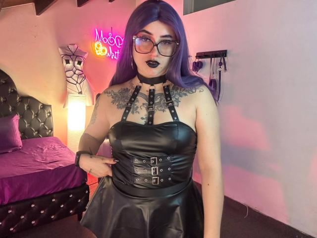 LilithDark69 met paars haar, een zwarte leren jurk met bandjes en gespen, tatoeages op de borst, zwarte lippenstift, een bril en een zwarte choker, staand in een zwak verlichte kamer..