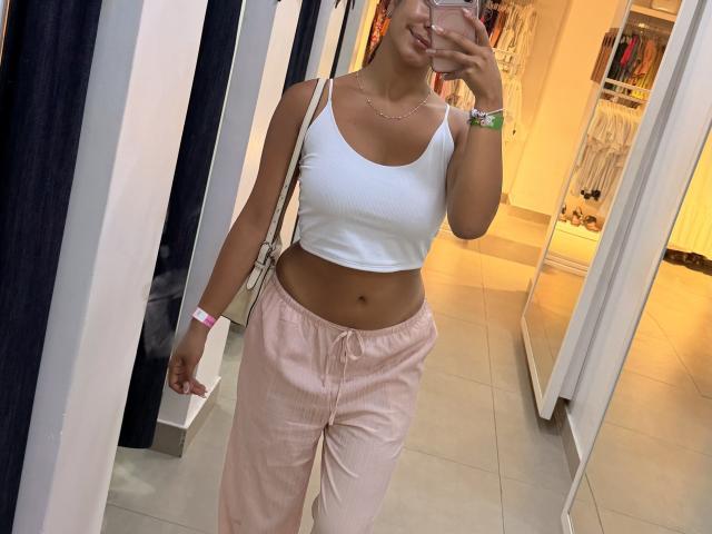 SarahPrincess with tan skin, white sports bra, pink pants, visible abs, holding phone, wearing pink wristband, necklace, standing in store, smiling, showing her tits and ass.