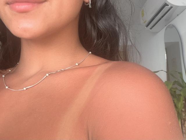 SarahPrincess's tan lines on her bare shoulders, wearing a delicate chain necklace, with her lips slightly smiling, dark wavy hair, and a small earring. Her skin is smooth.