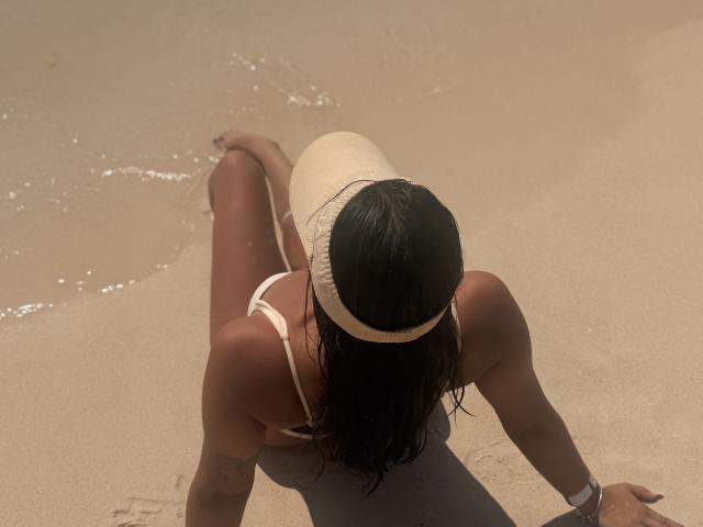 SarahPrincess with wet brown hair, wearing a white one-piece and a white headband, sitting on sandy beach, legs in shallow water, arms by her sides, back to camera.