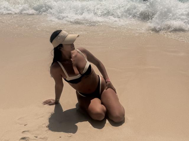 SarahPrincess kneeling on sandy beach, wearing black and white bikini, tan visor, dark hair, looking sideways, hands on wet sand, ocean waves in background, sunlight on her skin.