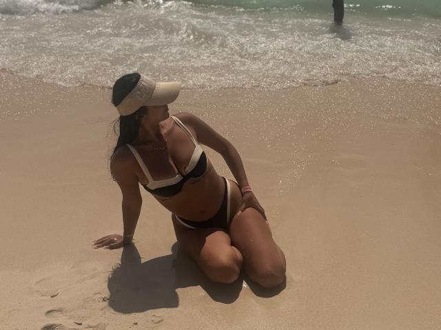 SarahPrincess kneels on sandy beach, wearing black and white bikini, sun visor, and pink wristband, with ocean waves in background, and a person standing in water.