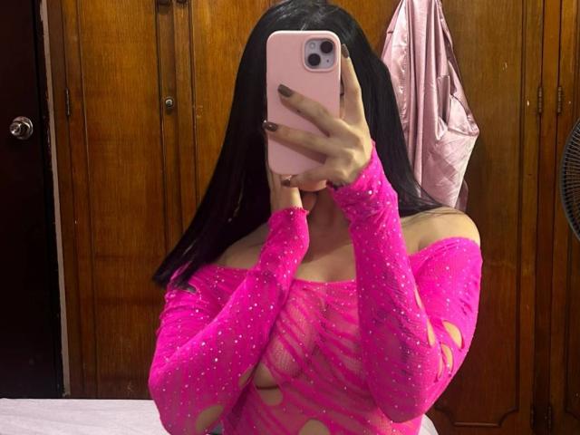 Katie Kingston mit langen, schwarzen Haaren, trägt ein durchsichtiges, pinkes Oberteil mit Ausschnitten, hält ein pinkes Telefon, bedeckt ihr Gesicht und zeigt ihre Brüste, in einem Zimmer mit Holzwänden..