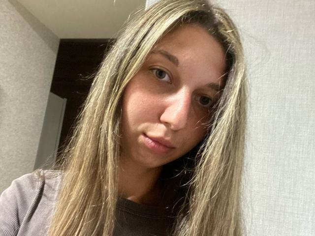 Sanella has long, straight blonde hair, light skin, and green eyes. She's wearing a brown ribbed shirt, with a neutral expression and slightly parted lips.