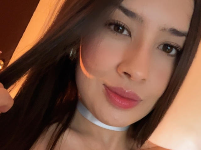 EmmaLombardi with long brown hair, wearing a white choker, has glossy pink lips and brown eyes, slightly smiling, with a warm light on her left cheek.