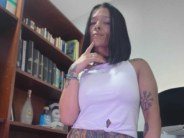 Amelie Blake met kort zwart kapsel, witte tanktop met een klein uitgesneden gedeelte, wijsvinger tegen haar kin, tatoeages op arm en been, staand voor een boekenkast, met een lichte glimlach..