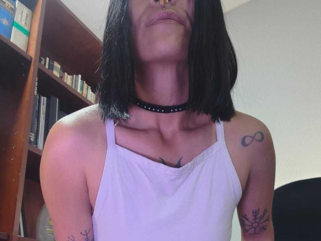 Amelie Blake, avec sa coupe de cheveux courte noire, son débardeur blanc, son collier noir, son piercing au nez, ses tatouages sur les épaules et ses lèvres légèrement entrouvertes, mettant en valeur sa petite poitrine et sa silhouette élancée..