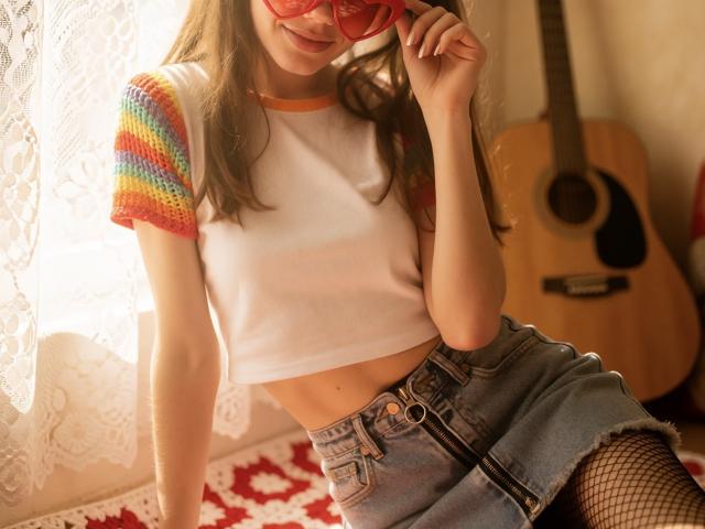 Euphorie, avec de petites seins, portant un haut en crochet multicolore, des lunettes en forme de cœur rouge, une jupe en jean et des bas résille, assise avec une guitare dans une pièce baignée de soleil..