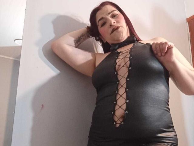 Molly Reed, met donkerrood haar, draagt een zwarte leren jurk met een veter sluiting aan de voorkant, waarbij haar borsten zichtbaar zijn, en staat tegen een witte muur, met haar hand op haar hoofd..