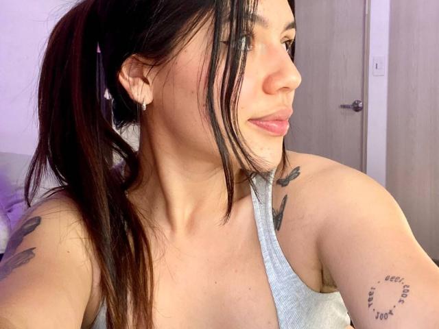SophiaMarx with long black ponytail, light gray tank top, small earrings, tattoos on shoulder and upper arm, smiling at camera, showing her tits and a cute face.