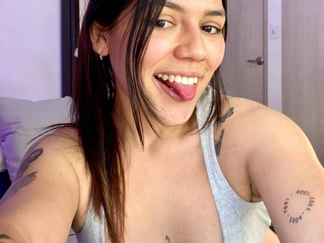SophiaMarx with long brown hair, smiling with tongue out, wearing a gray tank top, showing tattoos on her arm and side, with a cute face and small nose.